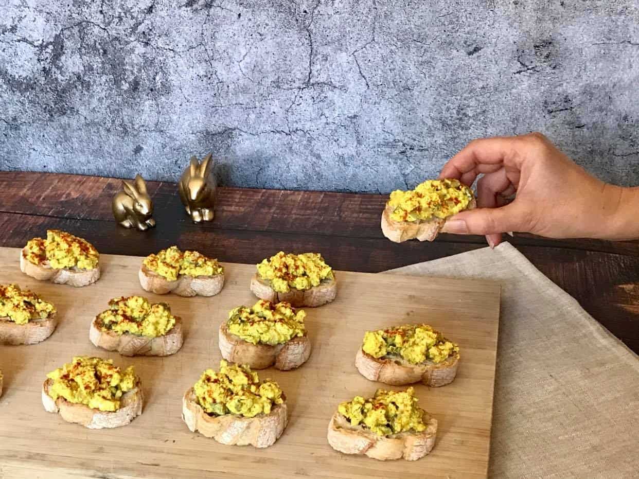 Vegan deviled egg crostini on a cutting board with a hand lifting one up.