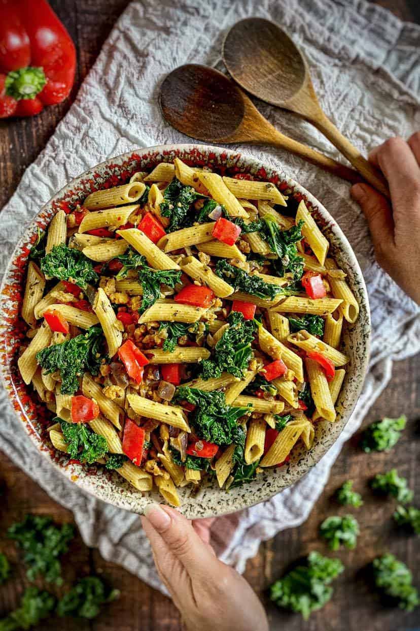 Vegan pasta with kale in bowl and hand holding two spoons.