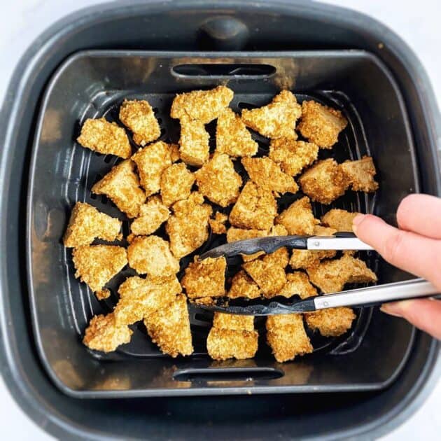 Hand flipping tofu nuggets in air fryer with tongs.