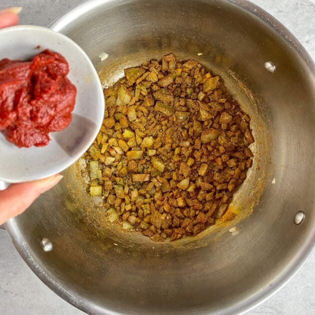 Onions and seasoning in a pot with hand holding a small bowl of tomato paste.