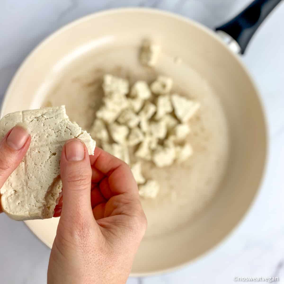 Hand breaking tofu into chunks over a skillet.