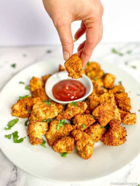 Air fryer tofu nuggets on a plate with a hand dipping one in ketchup.