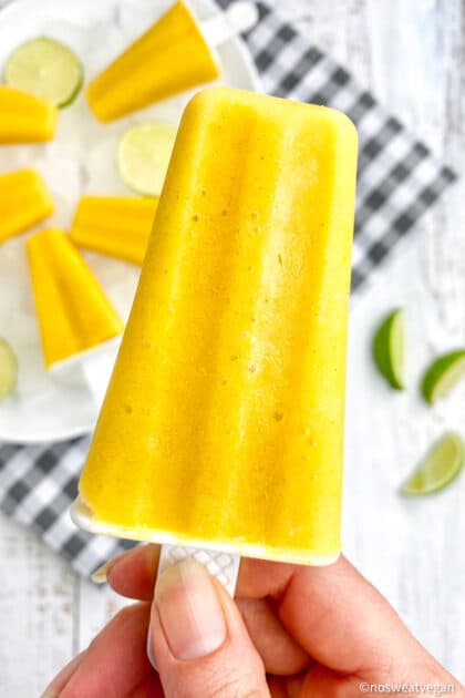 Mango popsicle held above a plate with 5 mango ice pops and limes.