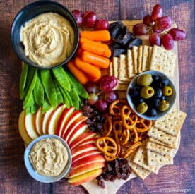 Vegan kids snack board with veggies, hummus, apple slices, cashew butter, graps, pretzels, whole wheat crackers, raisins, and dried apricots.