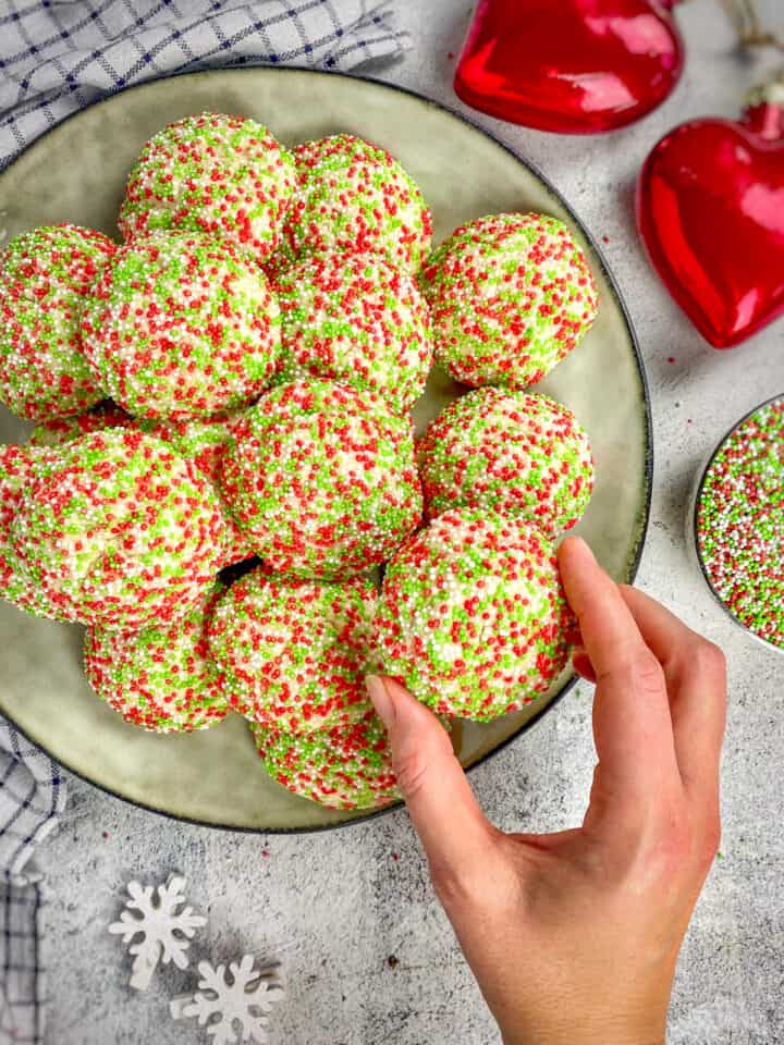 Christmas sprinkle cookies on a plate with a hand picking one up.