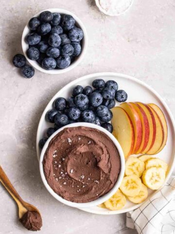 Chocolate hummus on a plate with blueberries, apples, and banana slices.