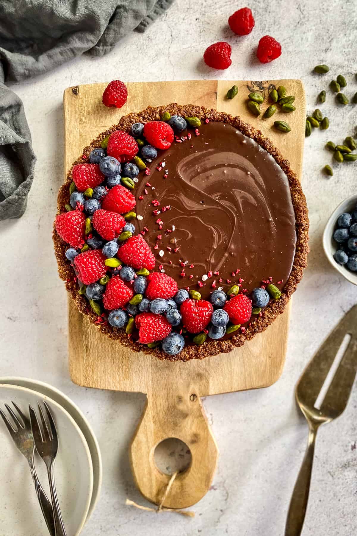 Chocolate tart on a cutting board and topped with raspberries, blueberries, and pistachios.