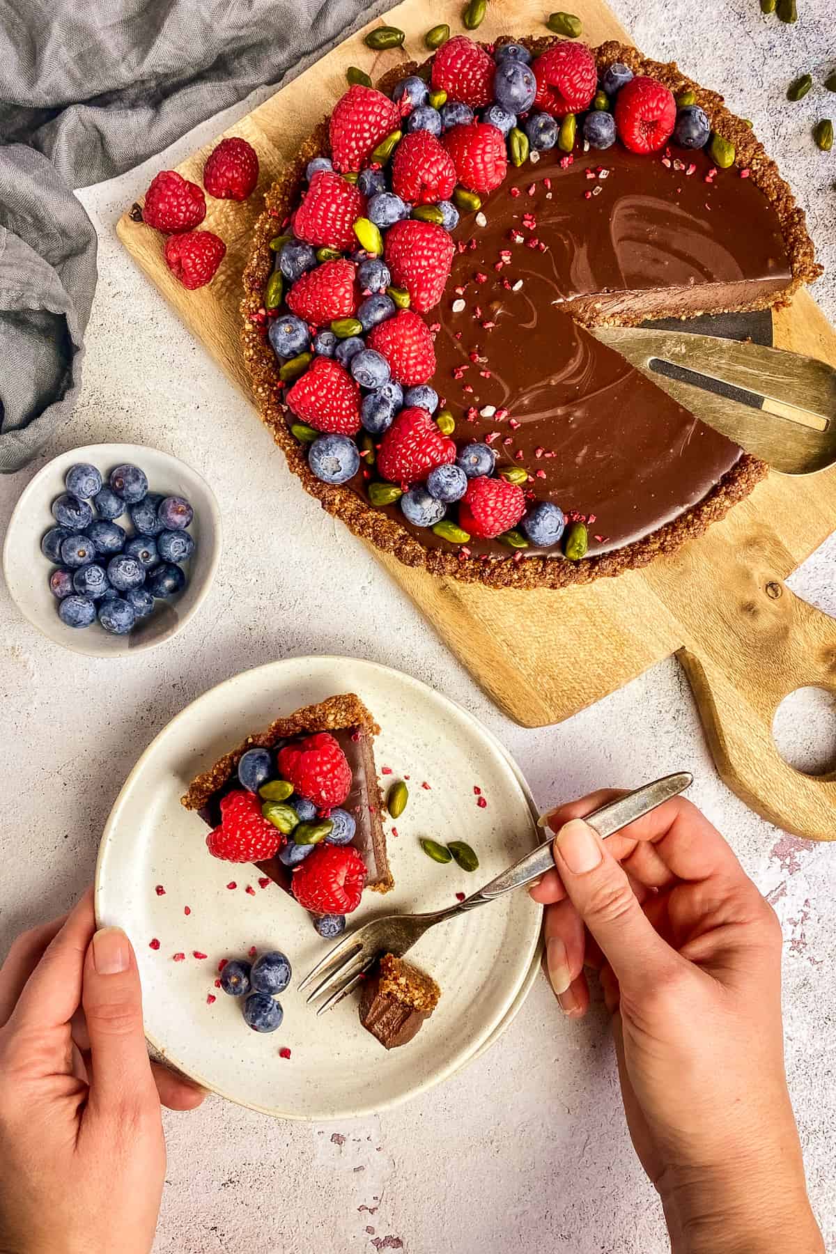 Vegan chocolate tart with berries on a cutting board next to a plate with a slice of the tart and two hands.