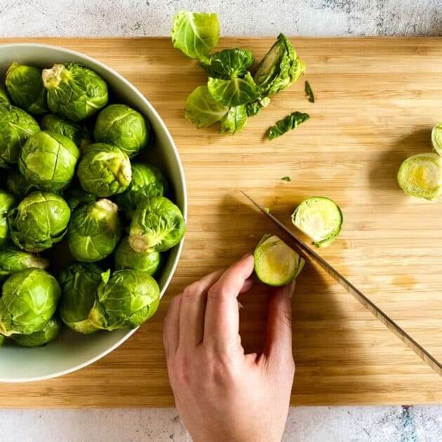 Knife cutting a brussel sprout in half on a cutting board next to a bowl of sprouts.