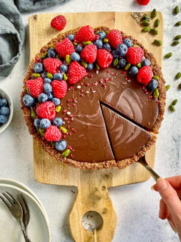 Vegan chocolate tart on a cutting board and topped with raspberries, blueberries, and pistachios.
