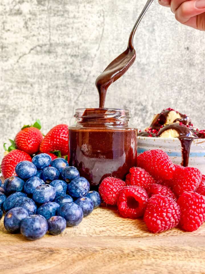 Hand lifting spoon of vegan chocolate sauce out of a jar surrounded by berries.