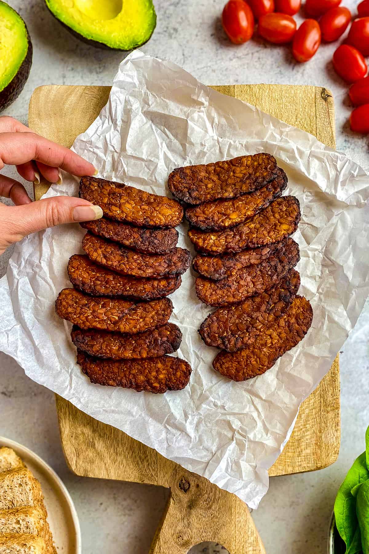 Tempeh bacon made in the air fryer on cutting board with hand taking one piece.