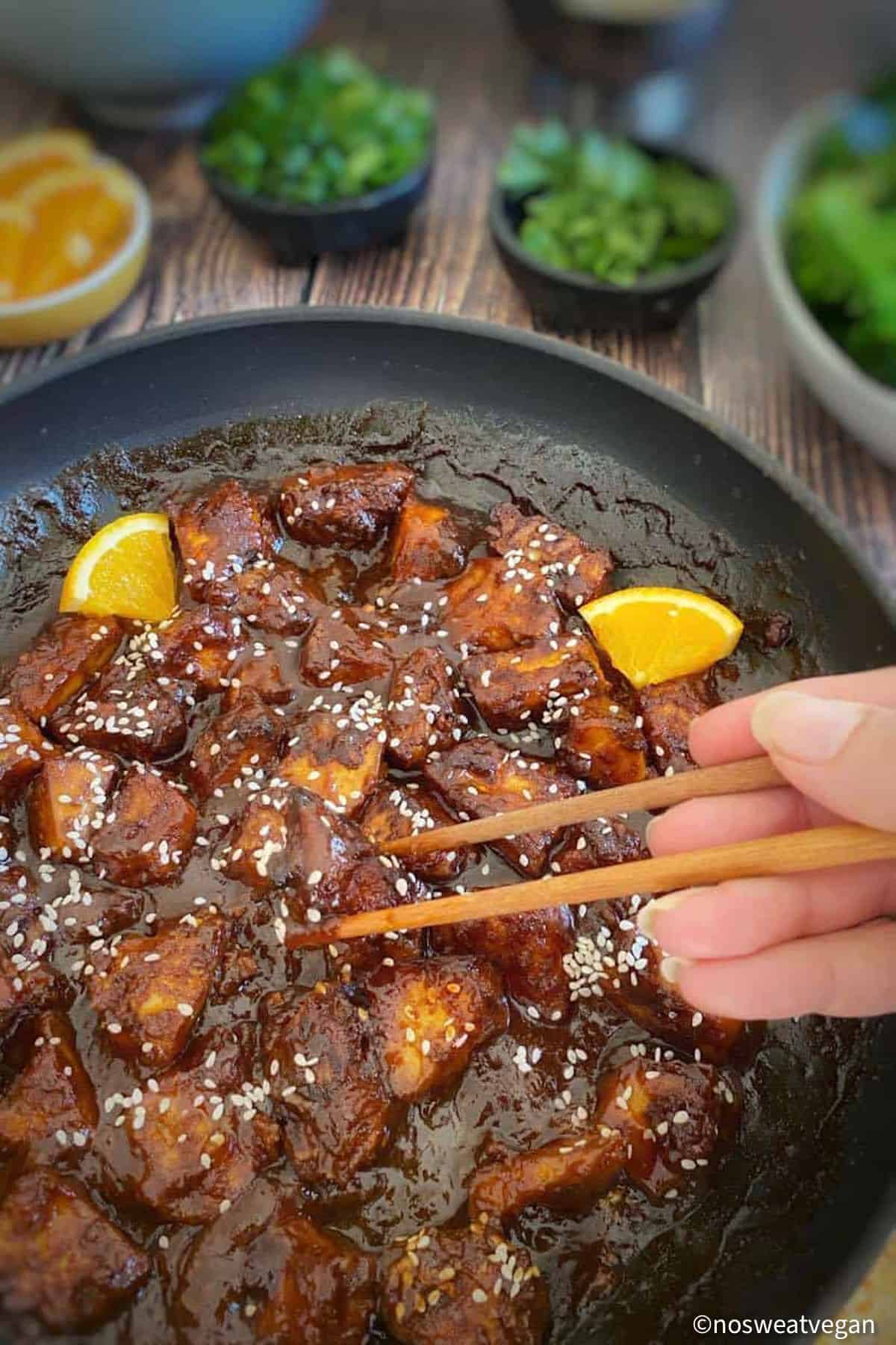 Skillet with vegan orange chicken and hand picking up a piece of tofu with chopsticks.