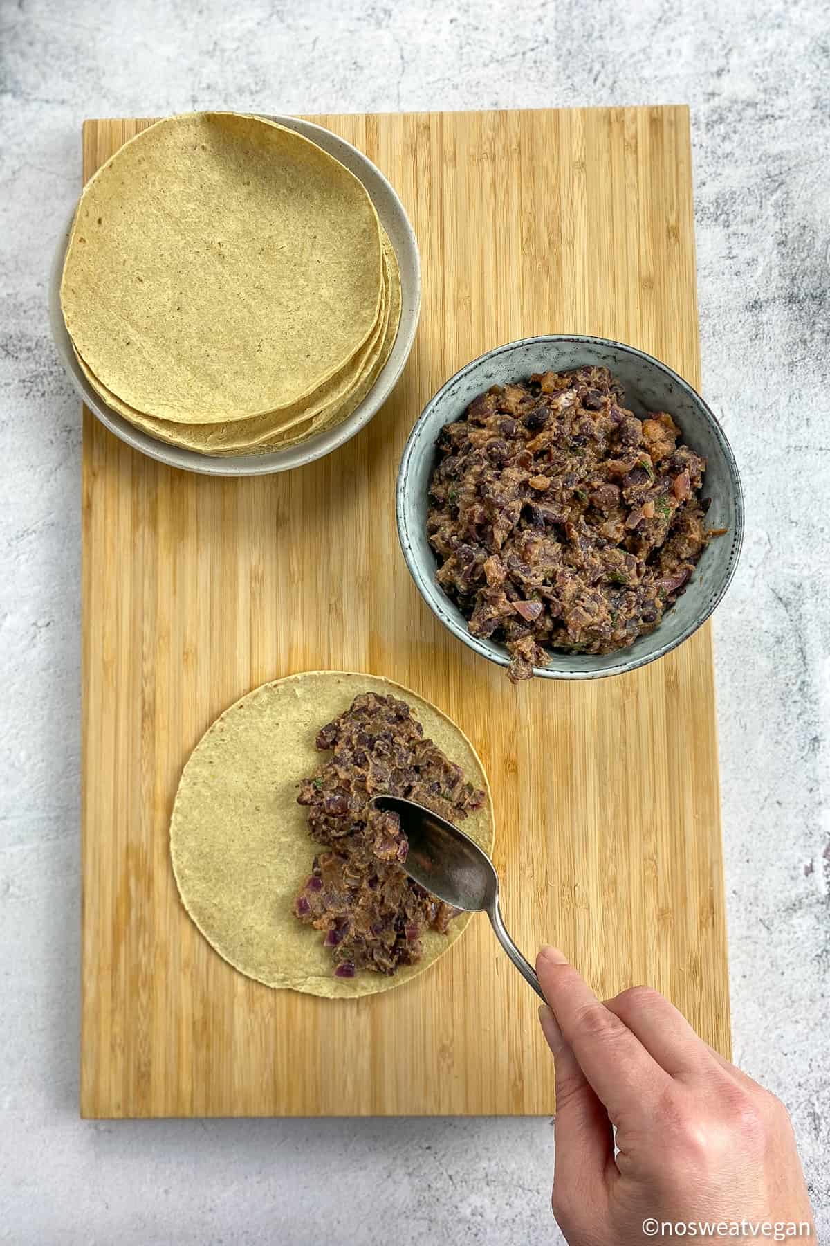 Taco shells, smashed black beans, and a hand adding the black bean filling to a taco.
