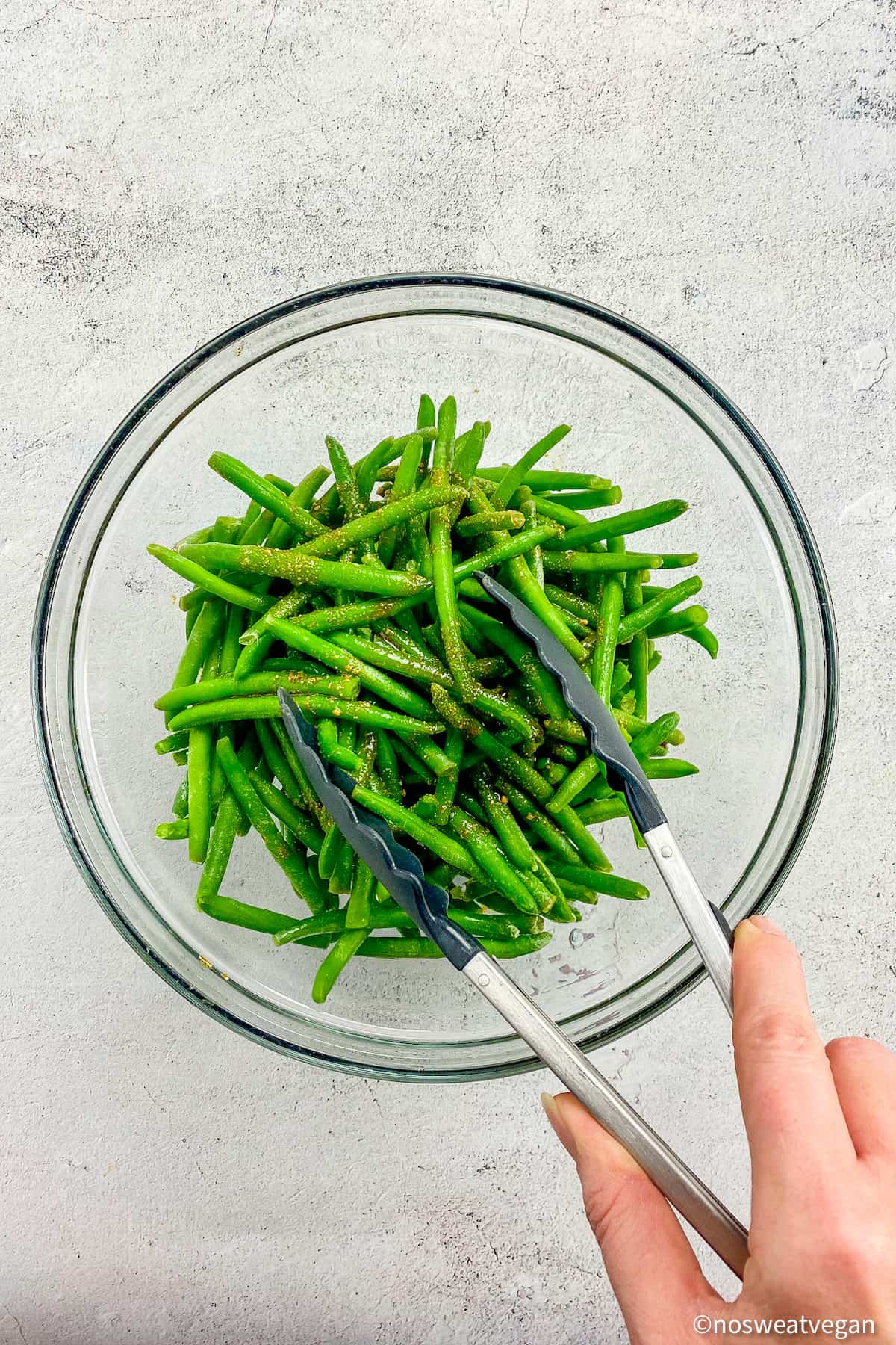 Frozen green beans in a bowl with seasoning and a hand holding tongs.